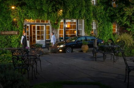A welcoming couple arrives at a charming ivy-covered hotel entrance, assisted by staff.