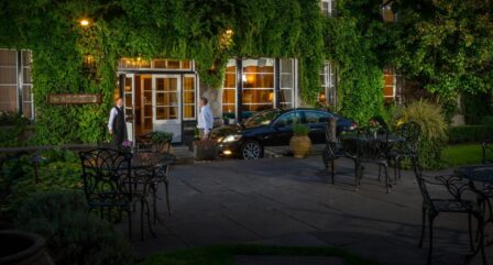 A welcoming couple arrives at a charming ivy-covered hotel entrance, assisted by staff.