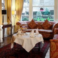 Cosy hotel lounge with a crackling fire, lavish sofas, and a table set for afternoon tea.