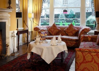 Cosy hotel lounge with a crackling fire, lavish sofas, and a table set for afternoon tea.