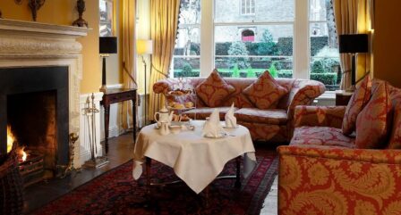 Cosy hotel lounge with a crackling fire, lavish sofas, and a table set for afternoon tea.