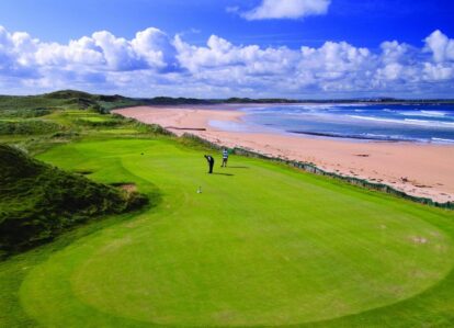 A couple enjoys golfing on a lush green course near a scenic beach under a blue sky.