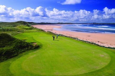 A couple enjoys golfing on a lush green course near a scenic beach under a blue sky.