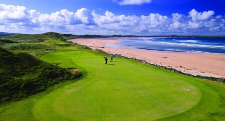 A couple enjoys golfing on a lush green course near a scenic beach under a blue sky.