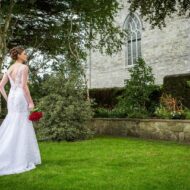 Bride in elegant dress holds red bouquet, standing in lush garden outside historic stone building.