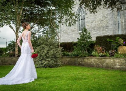 Bride in elegant dress holds red bouquet, standing in lush garden outside historic stone building.