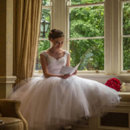 Bride reading a letter by a window in a cosy hotel lounge with lush garden view.