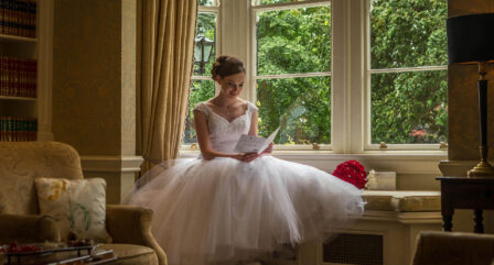 Bride reading a letter by a window in a cosy hotel lounge with lush garden view.