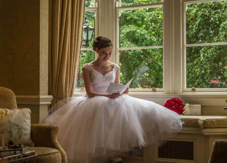 Bride reading a letter by a window in a cosy hotel lounge with lush garden view.