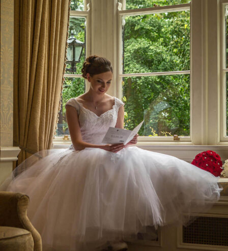 Bride reading a letter by a window in a cosy hotel lounge with lush garden view.