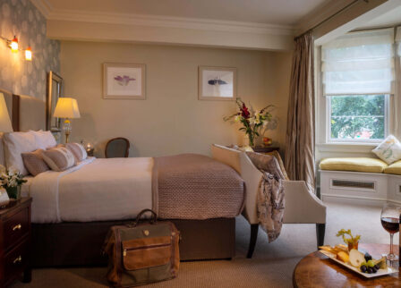 Cosy hotel room with plush bed, elegant decor, window seating, and wine on the table.