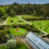 Aerial view of a lush garden with winding paths and a glasshouse, surrounded by trees.
