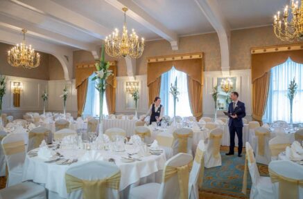 Elegant dining room set for an event, with staff preparing tables under grand chandeliers.