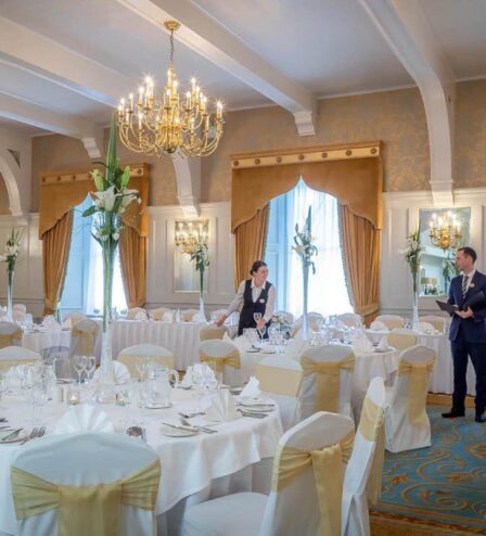 Elegant dining room set for an event, with staff preparing tables under grand chandeliers.